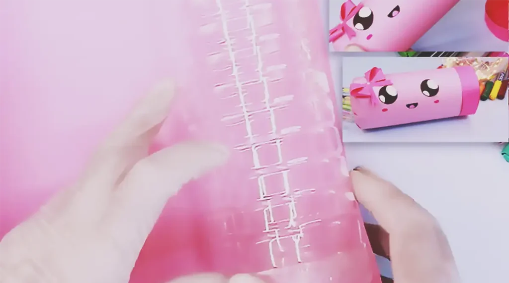 A cute pink pencil case made from a recycled plastic bottle, decorated with a kawaii face, bow, and filled with colorful markers for a DIY school supplies craft.