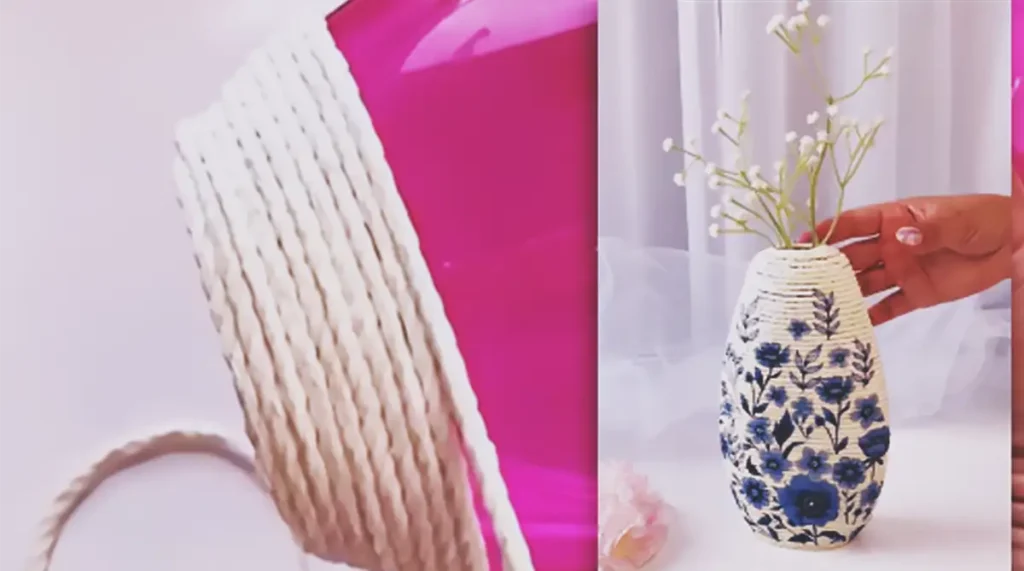 A handmade flower vase made from a recycled plastic bottle wrapped in cream rope and decorated with blue floral cutouts, holding small white flowers.
