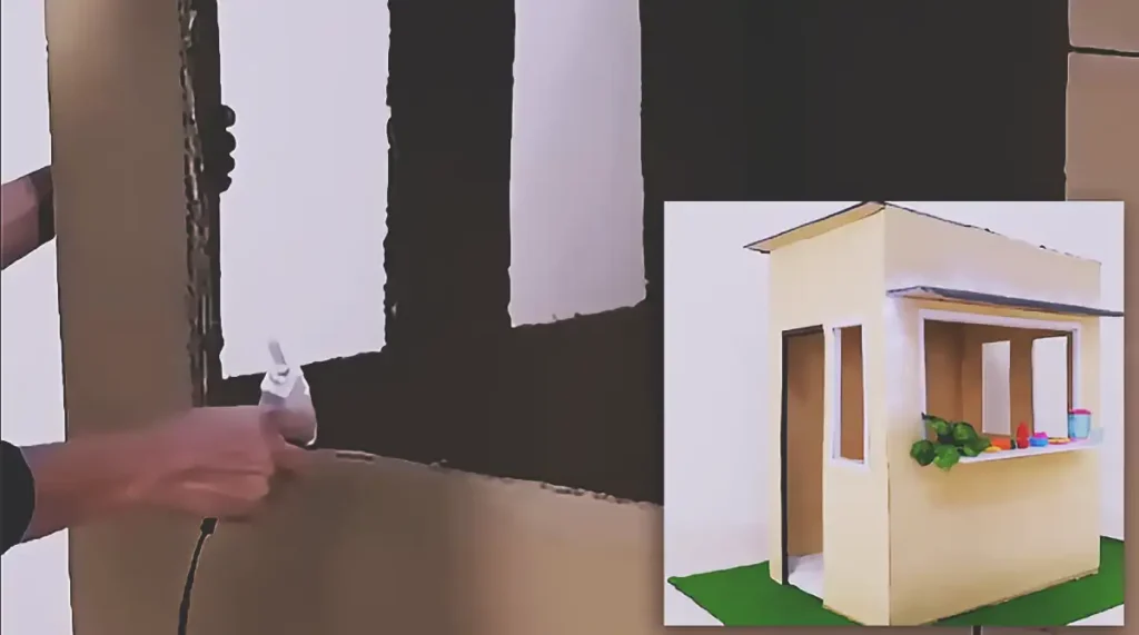 A finished child-sized playhouse made from large cardboard panels, featuring a wide serving window, cardboard counter, side doorway, narrow windows, dark roof awning, string lights, toy food, and a green floor mat.